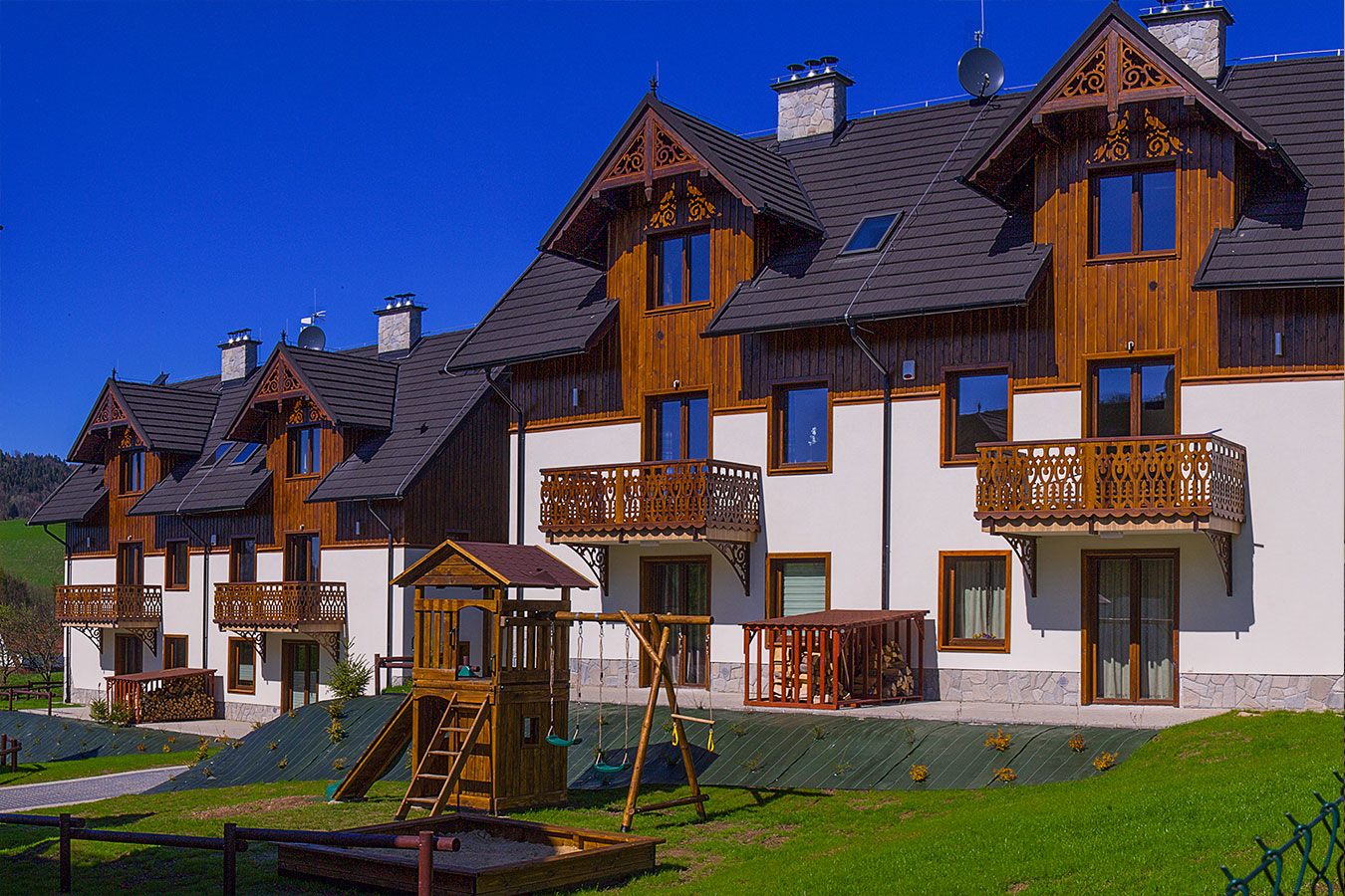 Zewnętrzna fasada apartamentów z tradycyjnymi drewnianymi zdobieniami oraz drewniany plac zabaw dla dzieci na zielonym trawniku