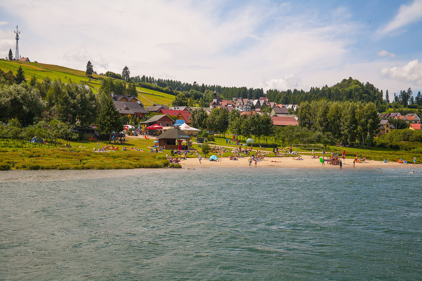 Słoneczna plaża i tereny rekreacyjne nad brzegiem Jeziora Czorsztyńskiego, w tle tradycyjna zabudowa i wzgórza Pienin